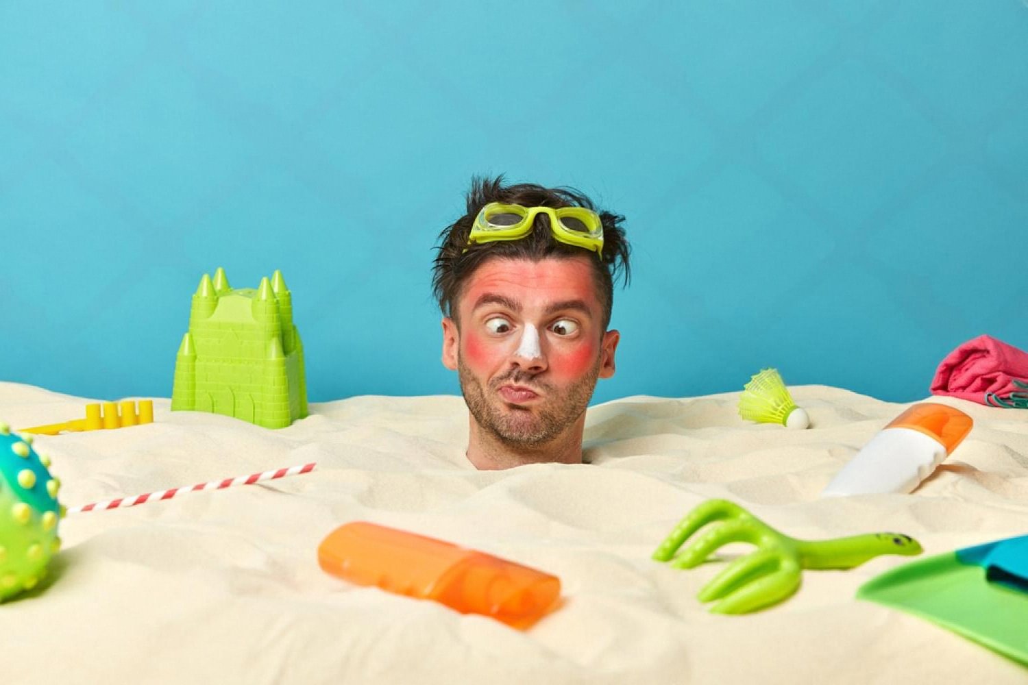 Man with severe sunburn on face poking out of the sand next to sunscreen bottles on a UAE beach